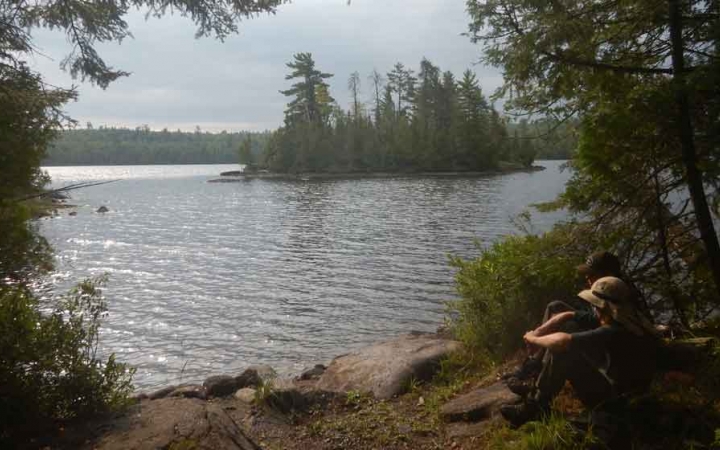 two people sit beside the shore of a lake and look toward a tree-lined island